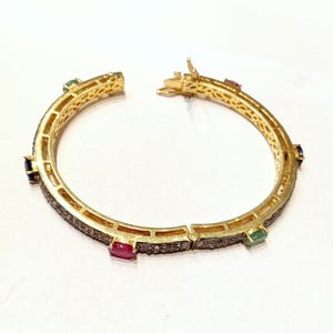 Könnte beinhalten: Ein goldenes Armreif-Armband mit einem diamantbesetzten Band und vier bunten Edelsteinen: einem Rubin, einem Smaragd und zwei Saphiren.