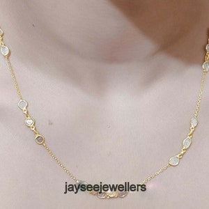 May include: A gold chain necklace featuring a row of clear, faceted stones. The stones are set in a bezel setting and are evenly spaced along the chain. The necklace is displayed on a model's neck.