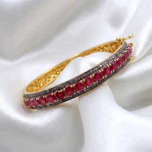 May include: A gold bangle bracelet featuring a row of red gemstones and a band of dark grey stones. The bracelet is displayed on a white, silky surface, accentuating its luxurious design.