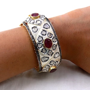 Może przedstawiać: Złota bransoletka typu bangle z geometrycznym wzorem ozdobiona czerwonymi rubinami i diamentami.
