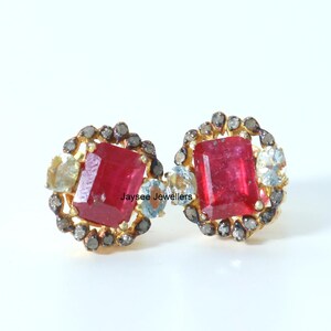 以下が含まれることがあります： 赤い正方形の宝石が小さなダイヤモンドで囲まれた、金色のイヤリングのペア。イヤリングは繊細で複雑なデザインです。