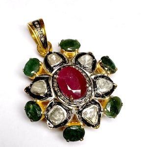 Puede incluir: Un colgante de oro con una piedra preciosa de rubí rojo rodeada de diamantes más pequeños y esmeraldas verdes. El colgante tiene la forma de una flor con una anilla en la parte superior.