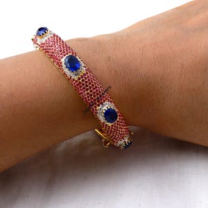 May include: A gold bangle bracelet with red and white stones and blue oval-shaped gemstones. The bracelet is on a person's wrist.