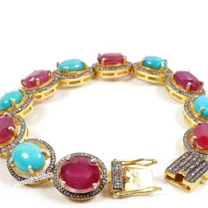 May include: A gold bracelet with alternating turquoise and ruby gemstones set in oval-shaped bezels. Each gemstone is surrounded by a halo of smaller diamonds. The bracelet has a gold clasp.