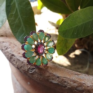 Può includere: Un anello d'oro con una grande pietra rossa circondata da pietre verdi più piccole e dettagli neri. L'anello ha la forma di un fiore.
