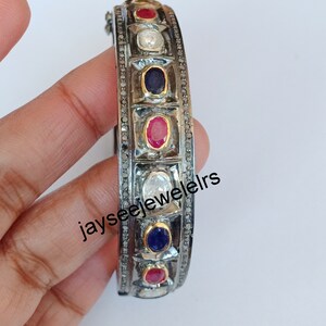 Op de afbeelding: Zilveren armband met een zwarte geoxideerde afwerking en een rij kleurrijke edelstenen. De armband heeft rode, blauwe en witte edelstenen die in een zilveren frame zijn gezet. De armband is versierd met kleine diamanten.