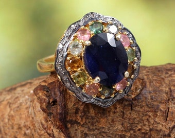 May include: A gold ring featuring a large blue sapphire surrounded by smaller gemstones in a variety of colours, including pink, green, and yellow. The ring is set in a silver band and has an ornate design.