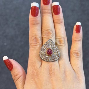 Puede incluir: Anillo de oro con forma de lágrima, con una piedra roja central y piedras claras circundantes. La mano muestra esmalte de uñas rojo brillante con puntas blancas.