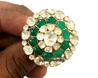 May include: A large, round ring with a gold setting and a design of green stones and white diamonds. The ring is set in a circular pattern with the green stones forming a ring around the centre of the ring. The ring is set on a finger.