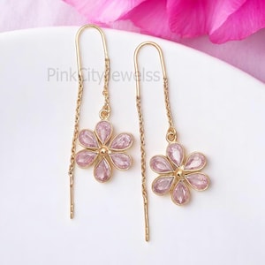 Puede incluir: Un par de pendientes de aro dorados con colgantes de flores rosas. Cada colgante de flor está hecho de pequeñas piedras preciosas rosas.