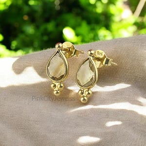 Puede incluir: Un par de pendientes de gota de oro con piedras transparentes facetadas. Los pendientes tienen un pequeño diseño de bola en la parte inferior.