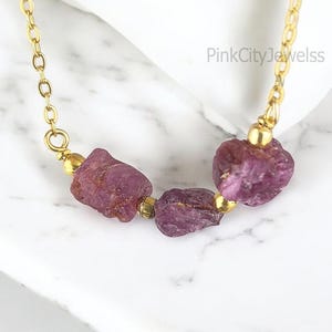 Könnte beinhalten: Eine goldene Kettenkette mit drei rosafarbenen Rubin-Edelsteinkugeln. Die Kette liegt auf einer weißen Marmorfläche. PinkCityJewels ist im Hintergrund geschrieben.