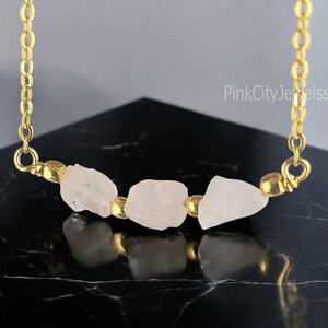 May include: A gold chain necklace with three raw rose quartz stones connected by gold wire. The stones are a light pink colour and are arranged in a row.