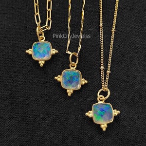 May include: Three gold necklaces with square pendants. Each pendant has a blue and green iridescent stone set in a gold frame with decorative accents. The necklaces hang against a black background.