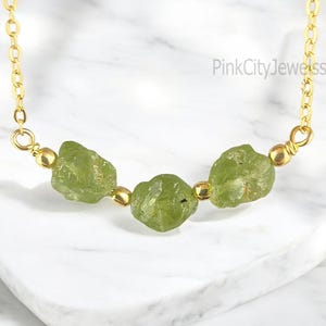 Könnte beinhalten: Eine goldene Kettenkette mit drei rohen grünen Peridot-Steinen. Die Steine sind unregelmäßig geformt und haben eine natürliche, raue Textur. Die Kette ist zart und schlicht, perfekt für den Alltag.