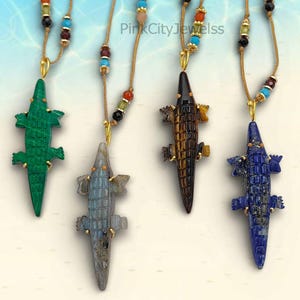 May include: Four gemstone alligator pendant necklaces. The pendants are green, grey, brown, and blue. Each alligator pendant is suspended from a cord necklace with colourful beads. The background is a light blue and white.