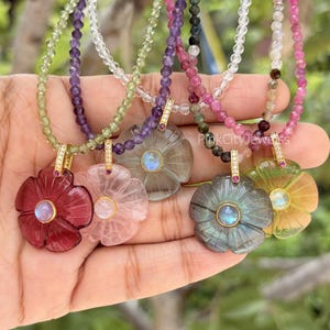 Puede incluir: Cinco collares con colgantes de piedras preciosas talladas en forma de flor en tonos rojo oscuro, rosa claro, gris translúcido, gris oscuro y amarillo. Cada colgante tiene una piedra central redonda y un engaste dorado con piedras claras.