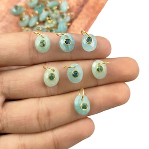 Puede incluir: Primer plano de ocho colgantes ovalados de piedra turquesa con anillas de salto doradas y pequeñas piedras verdes en el centro. Los colgantes se sostienen en una mano.