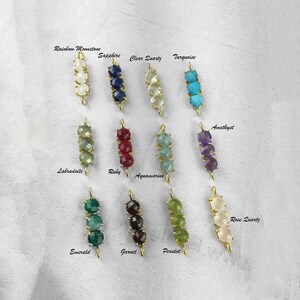 May include: A set of 12 gold charm pendants with different colored gemstones. Each pendant features three gemstones in a row. The gemstones include rainbow moonstone, sapphire, clear quartz, turquoise, labradoite, ruby, aquamarine, amethyst, emerald, garnet, peridot, and rose quartz.