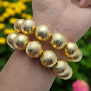 May include: Two gold-colored beaded bracelets. Each bracelet features large, textured, round beads. The bracelets are worn on a wrist, with a blurred background of green and yellow foliage. The text "PinkCityJewelss" is visible.