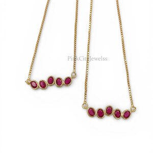 May include: Two gold necklaces, each featuring a row of five oval-cut, deep red gemstones. The gemstones are set in a gold bezel, and the necklaces hang from delicate gold chains. The background is plain white.