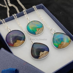 May include: Four silver necklaces with iridescent pendants in a blue box. The pendants have a wood and resin design, with colors ranging from blue to green. The necklaces are displayed in a blue jewelry box.