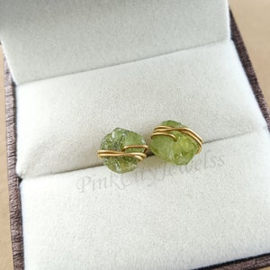 May include: A pair of peridot gemstone stud earrings. Each raw, light green peridot is wrapped with gold-coloured wire. The earrings are displayed inside a light grey jewellery box with a soft, velvety interior.
