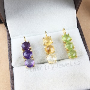 Könnte beinhalten: Drei goldene Anhänger, jeder mit drei Edelsteinperlen. Die Anhänger befinden sich in einer samtenen Box. Die Edelsteine sind Amethyst, Citrin und Peridot.