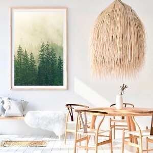 May include: A framed print of a misty forest scene with tall green trees. The print is hanging on a white wall above a wooden bench with a white faux fur throw. A large, natural woven pendant light hangs above a wooden dining table with four chairs.