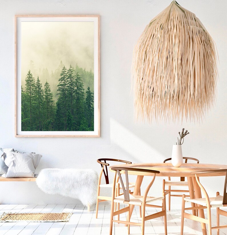 May include: A framed print of a misty forest scene with tall green trees. The print is hanging on a white wall above a wooden bench with a white faux fur throw. A large, natural woven pendant light hangs above a wooden dining table with four chairs.