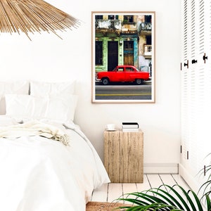 May include: A red vintage car parked in front of a colorful building with peeling paint. The car is in focus, while the building is slightly blurred in the background.