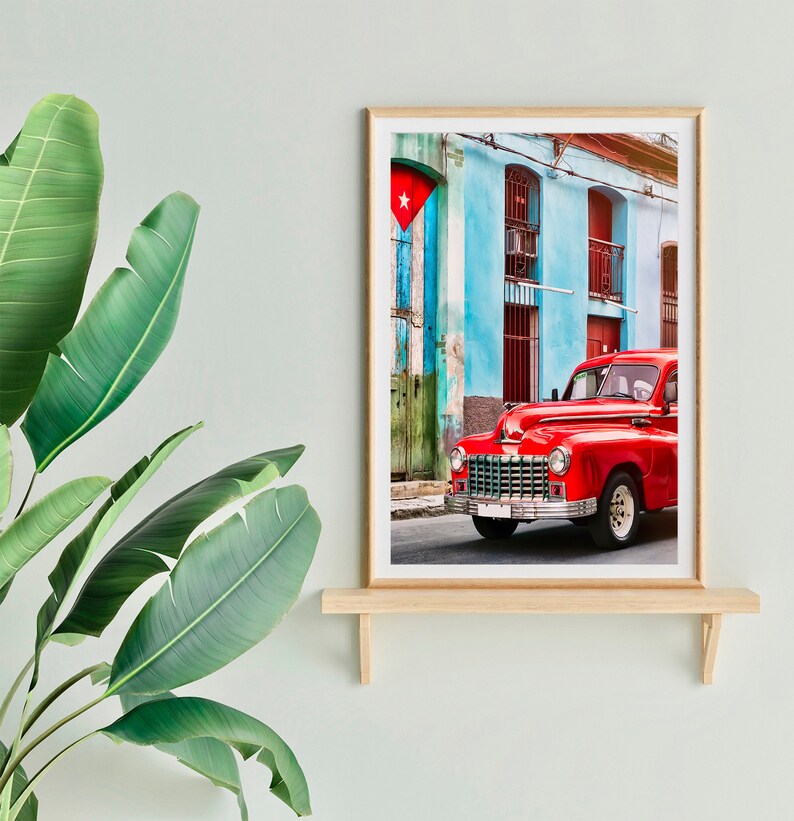 May include: A vintage red car parked in front of a blue and green building with a Cuban flag hanging on the wall. The car has a white star on the front grill and a white license plate.