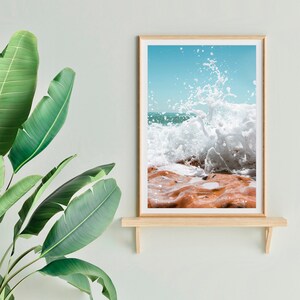 May include: A framed print depicting a crashing wave. The image shows a turquoise sky, white water, and orange-brown rocks. The frame is light wood, and the print is displayed on a wooden shelf.