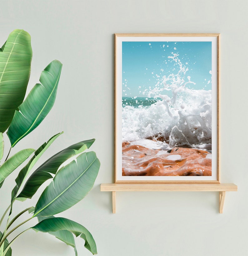 May include: A framed print depicting a crashing wave. The image shows a turquoise sky, white water, and orange-brown rocks. The frame is light wood, and the print is displayed on a wooden shelf.