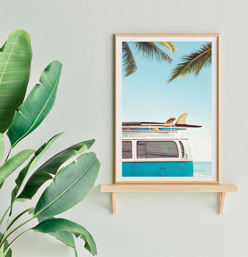 May include: A framed print of a blue vintage van with surfboards on the roof, parked on a beach with palm trees in the background.