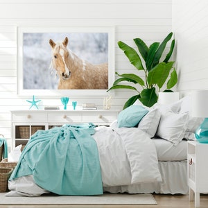May include: A bedroom scene with a large framed print of a horse. The room features a white dresser with decorative items, a bed with white linens, and a turquoise throw blanket. A large green plant adds a natural element.