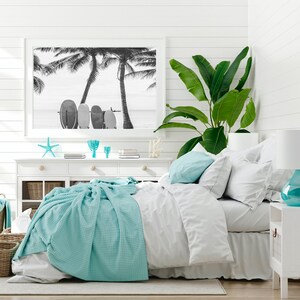 K&ouml;nnte beinhalten: Ein wei&szlig;es Schlafzimmer mit einem Bett, einer wei&szlig;en Kommode und einer Pflanze. Das Bett hat einen wei&szlig;en Bettbezug und eine hellblaue Decke. &Uuml;ber der Kommode h&auml;ngt ein Schwarzwei&szlig;foto von Palmen und Surfbrettern.