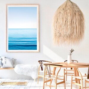 May include: A white, fluffy rug sits in front of a wooden bench with a white throw pillow. A framed photograph of a blue ocean hangs on the wall above the bench. A large, woven, straw pendant light hangs from the ceiling above a wooden dining table with four chairs.