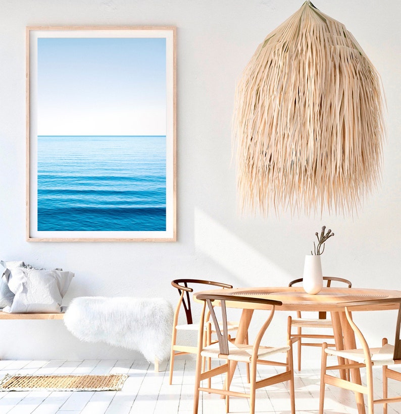 May include: A white, fluffy rug sits in front of a wooden bench with a white throw pillow. A framed photograph of a blue ocean hangs on the wall above the bench. A large, woven, straw pendant light hangs from the ceiling above a wooden dining table with four chairs.