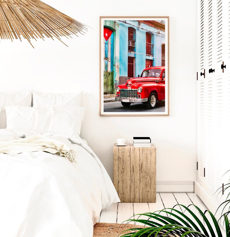 May include: A red vintage car parked in front of a blue and green building with a Cuban flag hanging from the building. The car has chrome accents and whitewall tires.