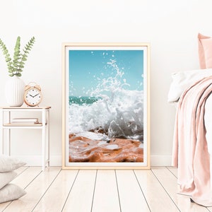 May include: Framed print depicting a powerful ocean wave breaking over reddish-brown rocks against a clear, light blue sky. The artwork is presented in a light wood frame, creating a coastal atmosphere.