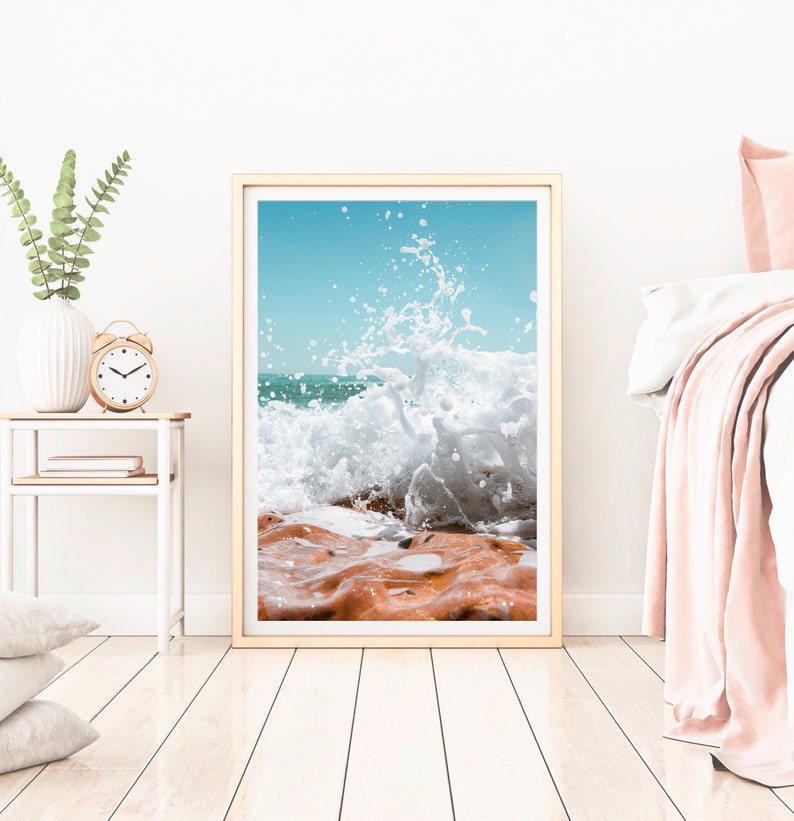 May include: Framed print depicting a powerful ocean wave breaking over reddish-brown rocks against a clear, light blue sky. The artwork is presented in a light wood frame, creating a coastal atmosphere.