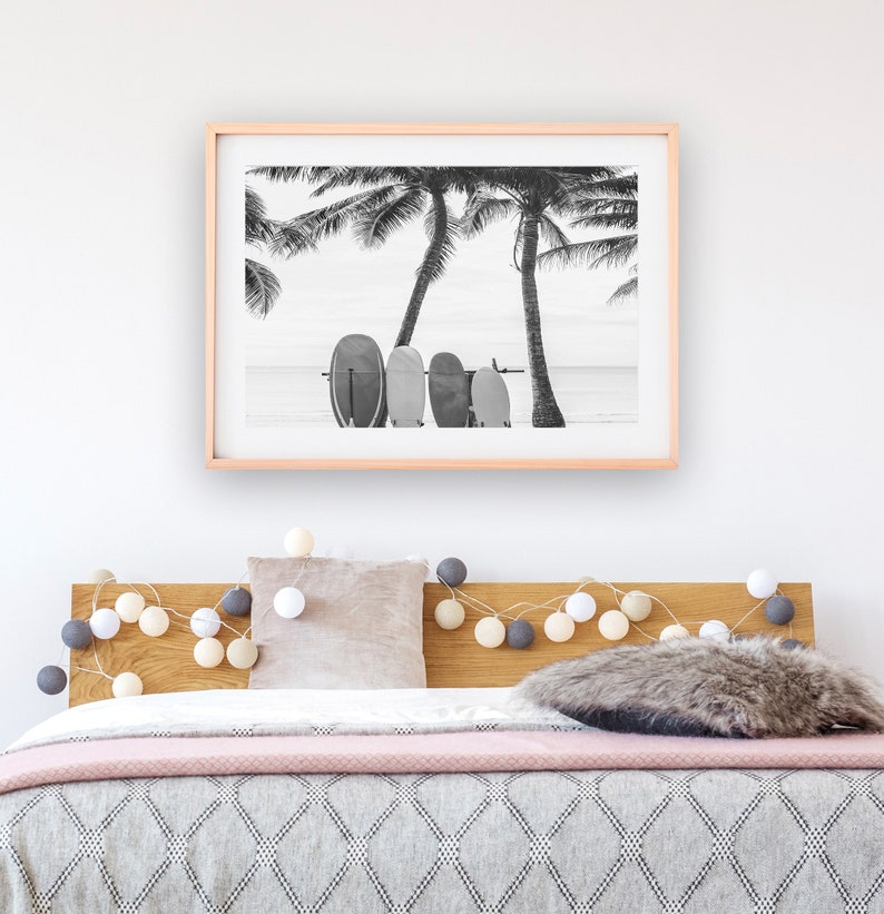 K&ouml;nnte beinhalten: Schwarzwei&szlig;foto von Palmen und Surfbrettern an einem Strand. Das Foto ist gerahmt und h&auml;ngt &uuml;ber einem Bett mit einem Holz-Kopfteil und einer rosa-grauen Decke.