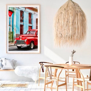 May include: A red vintage car parked in front of a colorful building with a Cuban flag hanging on the side. The car has a chrome bumper and whitewall tires. The building has blue and green walls with white trim.