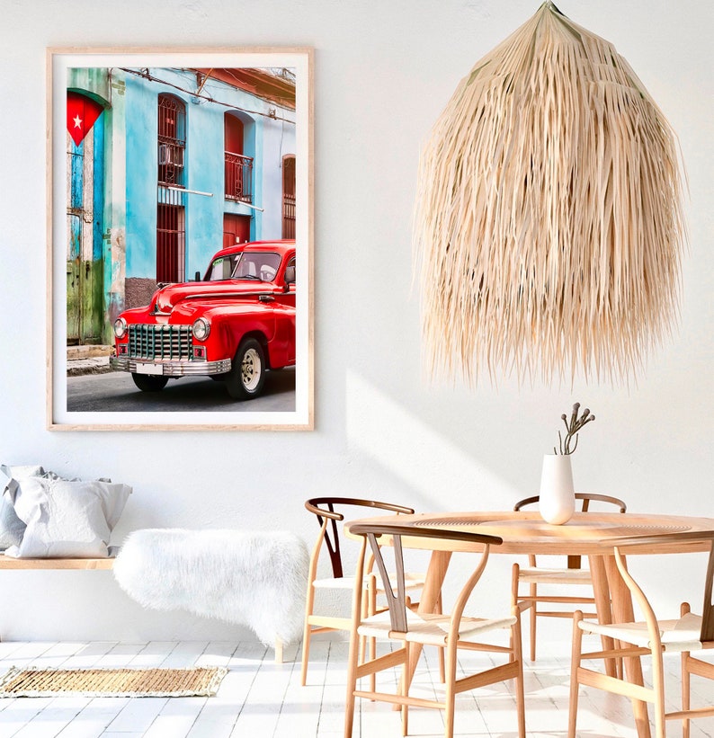 May include: A red vintage car parked in front of a colorful building with a Cuban flag hanging on the side. The car has a chrome bumper and whitewall tires. The building has blue and green walls with white trim.