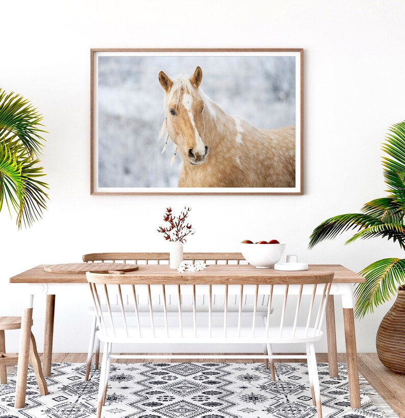 May include: A framed print featuring a close-up of a palomino horse with a light brown coat and white mane. The horse has a serene expression and is adorned with decorative feathers. The artwork is displayed above a wooden dining table.