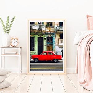 May include: A vintage red car parked in front of a colorful building with green, blue, and yellow accents. The building has multiple windows and balconies.