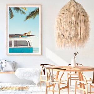 May include: A blue vintage van with surfboards on the roof parked on a beach with palm trees in the background. The van is in front of a white wall with a large framed print of the same scene.