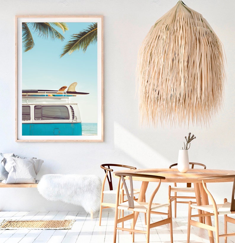 May include: A blue vintage van with surfboards on the roof parked on a beach with palm trees in the background. The van is in front of a white wall with a large framed print of the same scene.