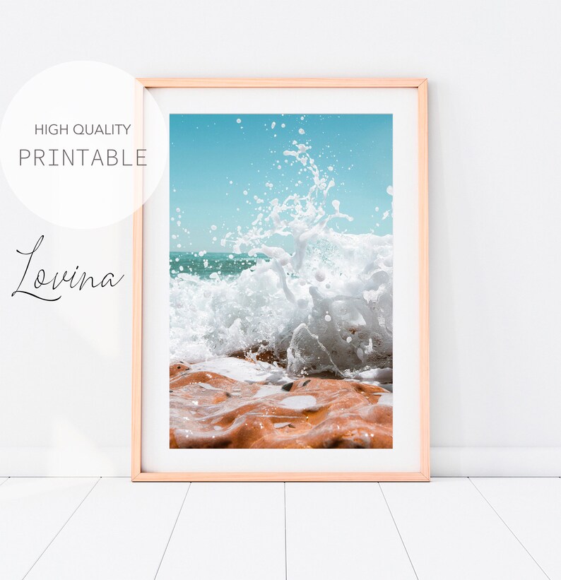 May include: Framed print depicting a dynamic ocean scene. A wave breaks over reddish-brown rocks, with white foam and a turquoise sea under a clear blue sky. The print is labeled "HIGH QUALITY PRINTABLE" and "Lovina".